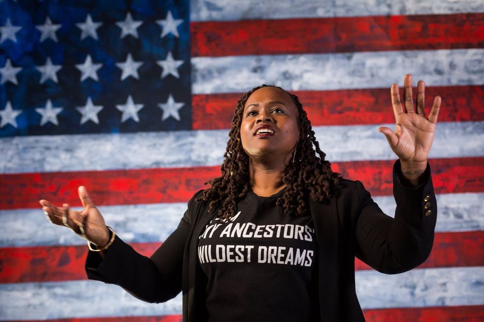 Photo of Jamilia Lyiscott standing in front of the American Flag wearing a black T-shirt that says I am my ancestors' wildest dreams