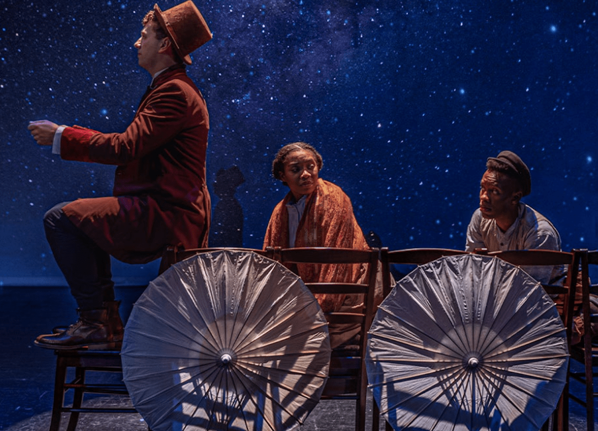 A scene from the production with actors in a makeshift wagon created with chairs and umbrellas. 