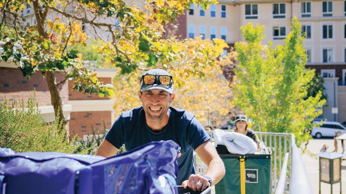 Smiling person pushing a green moving bin of belongings near campus apartments