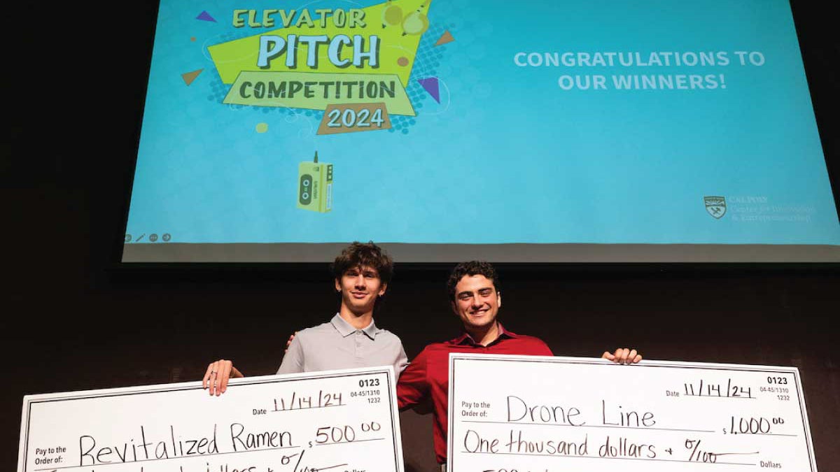 Two male Cal Poly students standing on a stage holding large checks for $500 and $1,000 for winning the 2024 Elevator Pitch Competition.