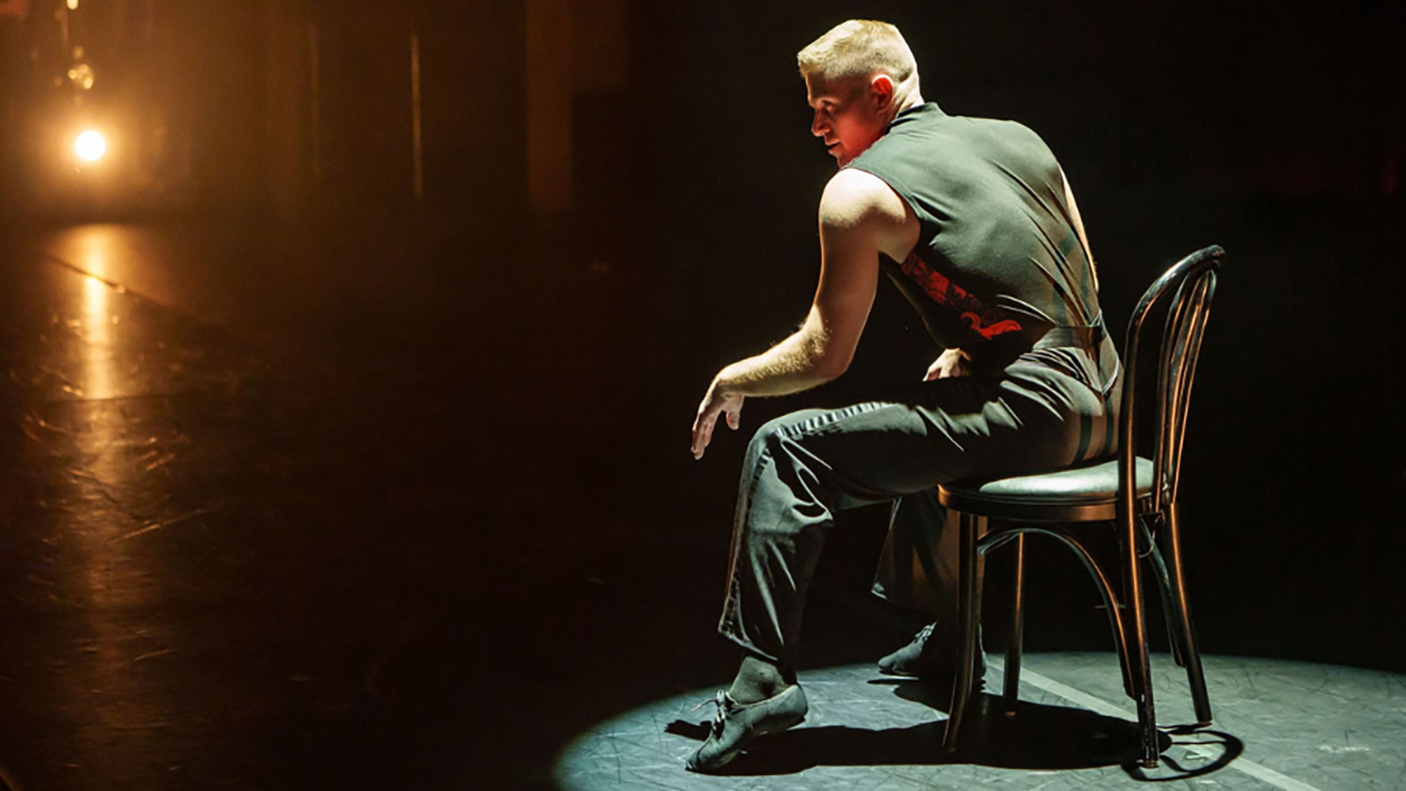 A man dressed in all black sitting on a chair and performing on stage.