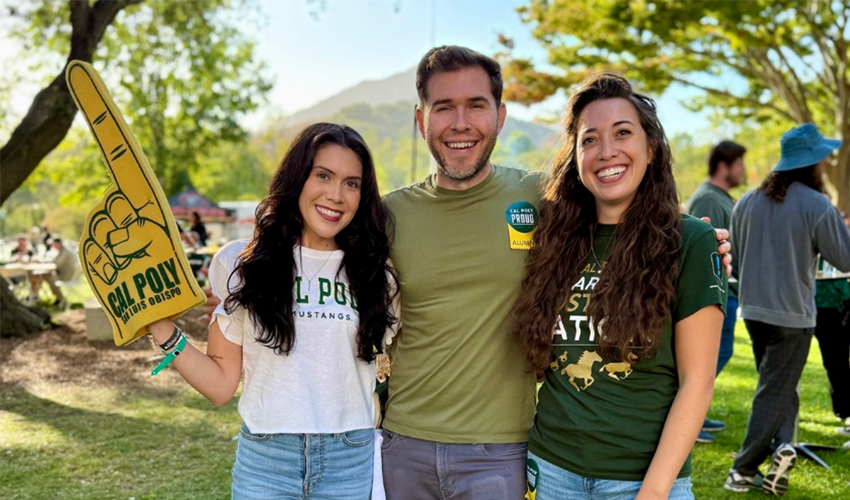 Three Cal Poly alumni smile outdoors, with one holding a large Cal Poly foam finger.