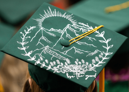 Aerial shot of green graduation cap with the words "be the good" painted on it.