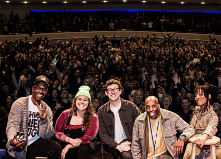 Comedians pose on stage with crowd in background.