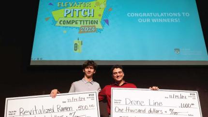 Two male Cal Poly students standing on a stage holding large checks for $500 and $1,000 for winning the 2024 Elevator Pitch Competition.
