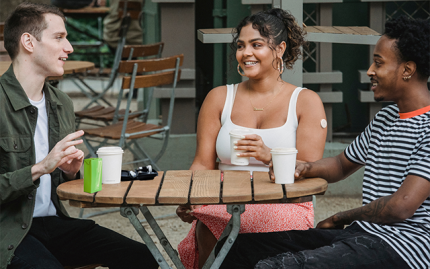 Three students sitting at a table with drinks and talking. One has a blood sugar monitor.