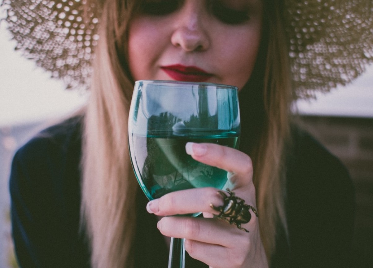 Woman with red lipstick and wearing a straw hat holds green tinted wine glass