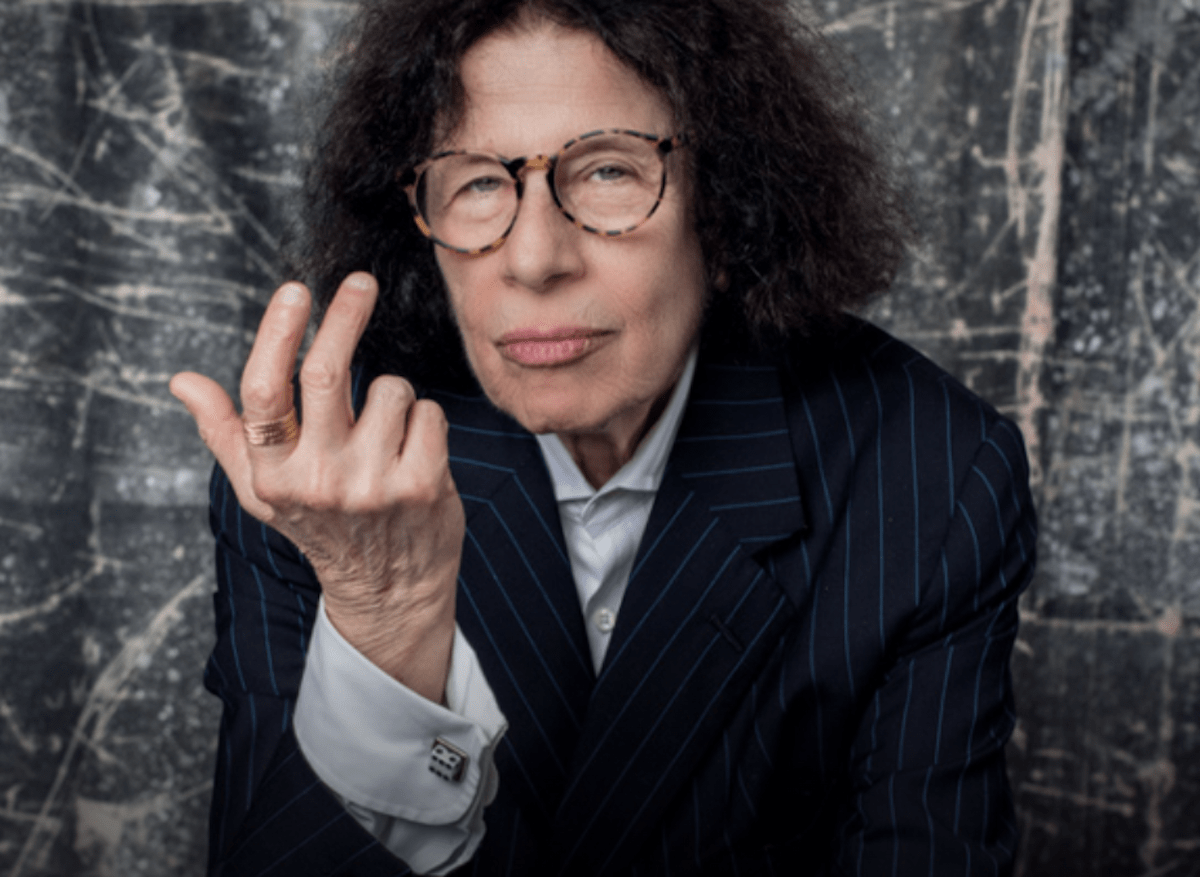 Photo of Fran Lebowitz sitting in a blue suite in front of a marbled gray wall. 