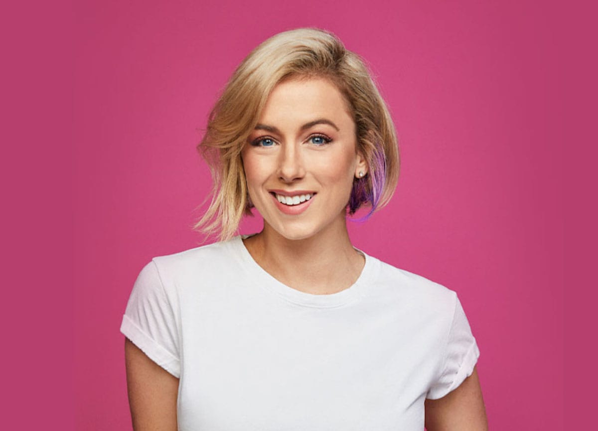 Photo of Iliza Schlesinger. She is wearing a white t-shirt. The background is pink.