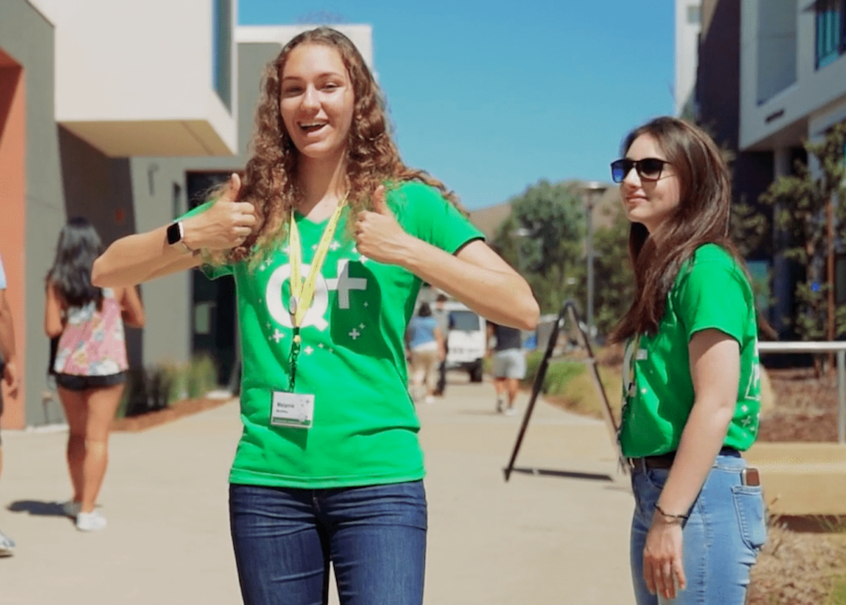 Photo of 2 Quarter Plus students. One is facing the camera giving a two-thumbs up and the other is standing to the side. Both are wearing green Quarter Plus t-shirts.